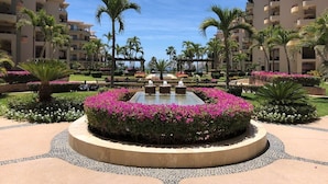 Un jardín paisajístico con una fuente central, rodeado de palmeras y plantas florecientes, y edificios de varios pisos al fondo.