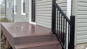 Une maison avec un revêtement extérieur gris, une terrasse en bois avec des marches et une rambarde en métal noir.