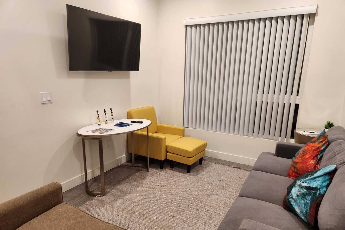 Un salón con un sofá gris, un sillón amarillo, una mesita blanca y un televisor montado en la pared.