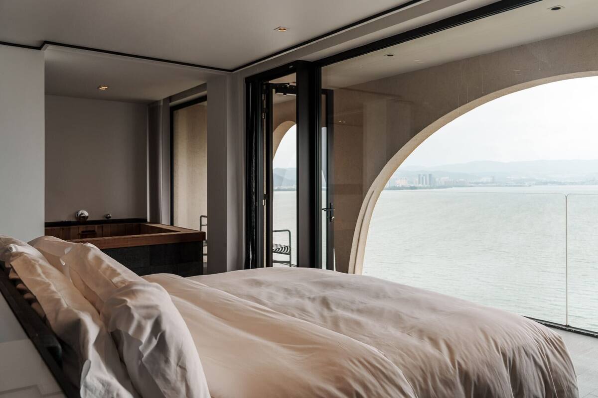 Ein modernes Schlafzimmer mit einem großen Bett, einer hölzernen Bank und Blick auf ein Gewässer durch ein großes Fenster.