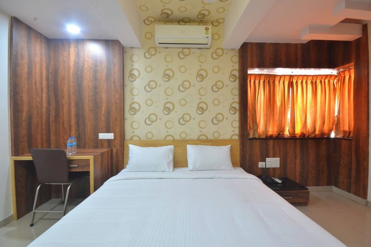 A hotel room with a bed, a desk with a chair, an air conditioner, and a window with curtains.