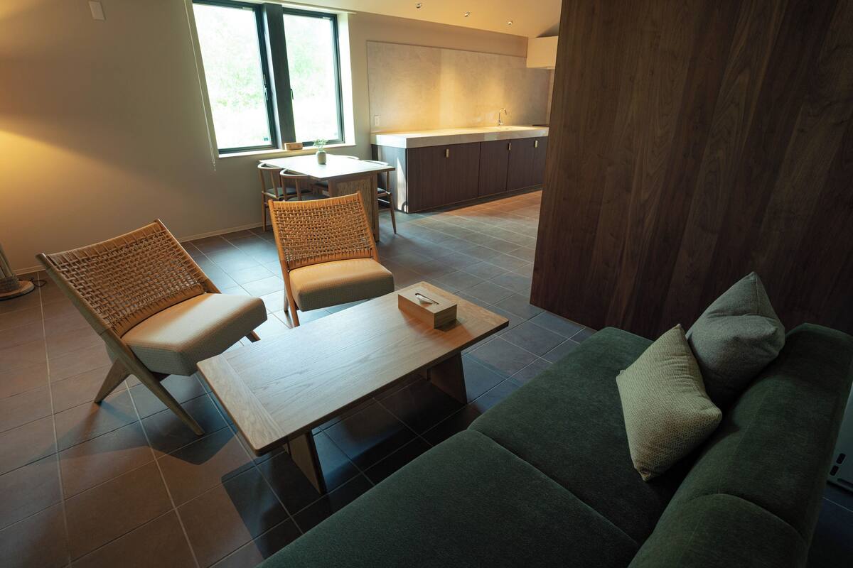A modern living room with a green sofa, two wicker chairs, a wooden coffee table, and a kitchen in the background.