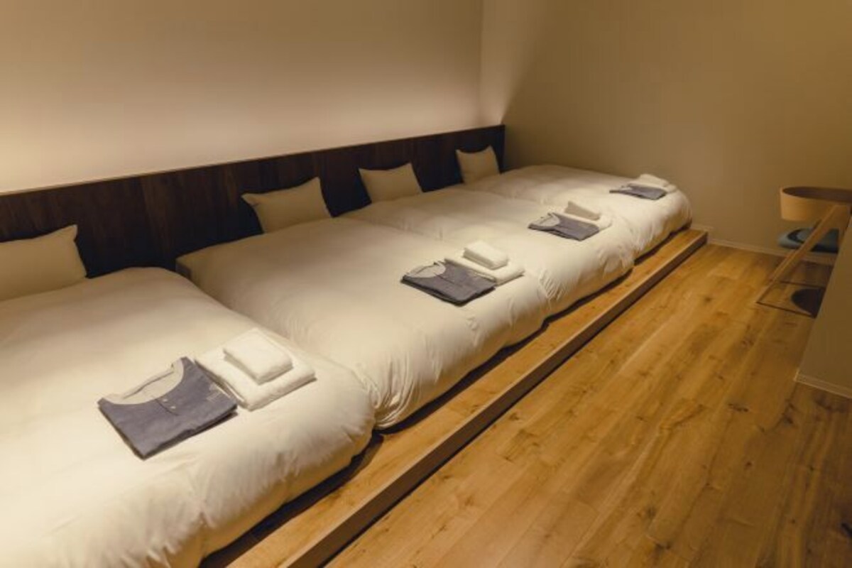 Cuatro camas individuales con estructura de madera y ropa de cama blanca, dispuestas en una habitaci ón estrecha con suelo de madera.