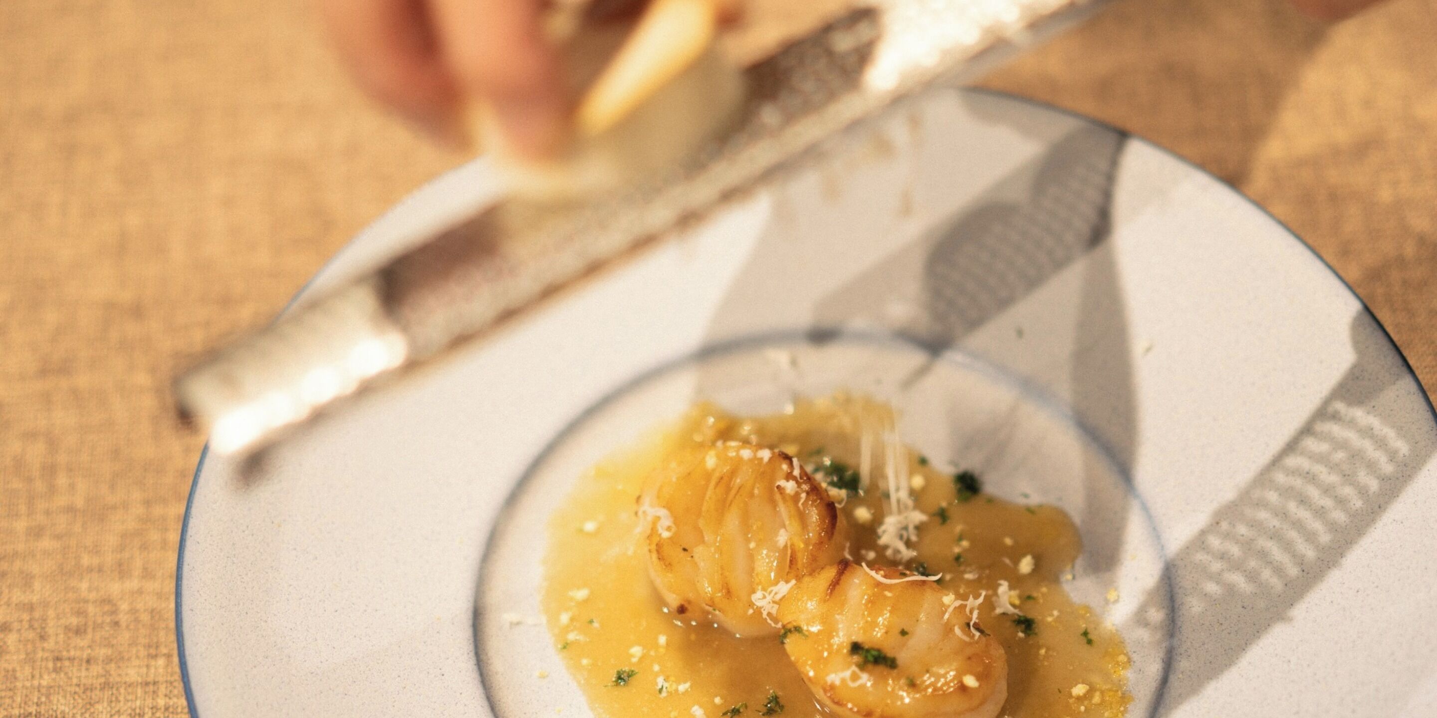 Un plato con un guiso de mariscos, posiblemente vieiras, adornado con hierbas y queso, sobre un mantel texturizado.