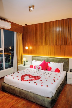 A neatly made bed with rose petals and a heart-shaped arrangement, a wooden headboard, a nightstand with a lamp, and a window with curtains. - Mhendo Hotel & Studio Suites (Burhanilkantha)