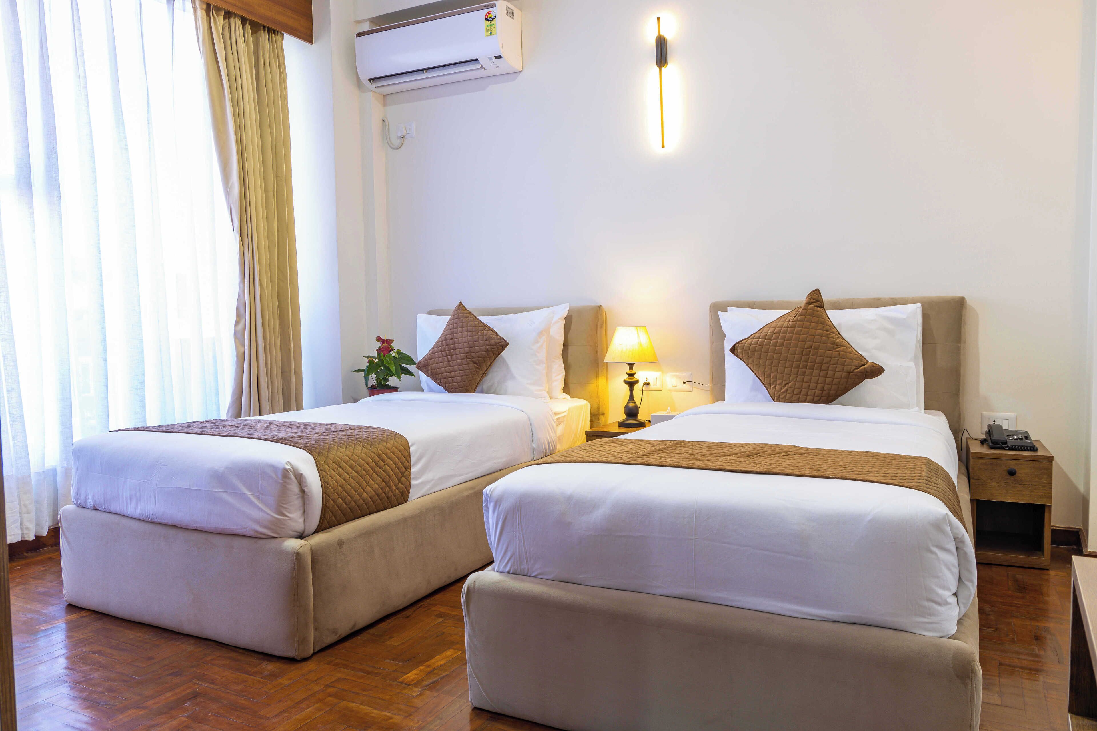 A hotel room with two beds, a bedside table with a lamp, a nightstand with a phone, and an air conditioning unit on the wall.