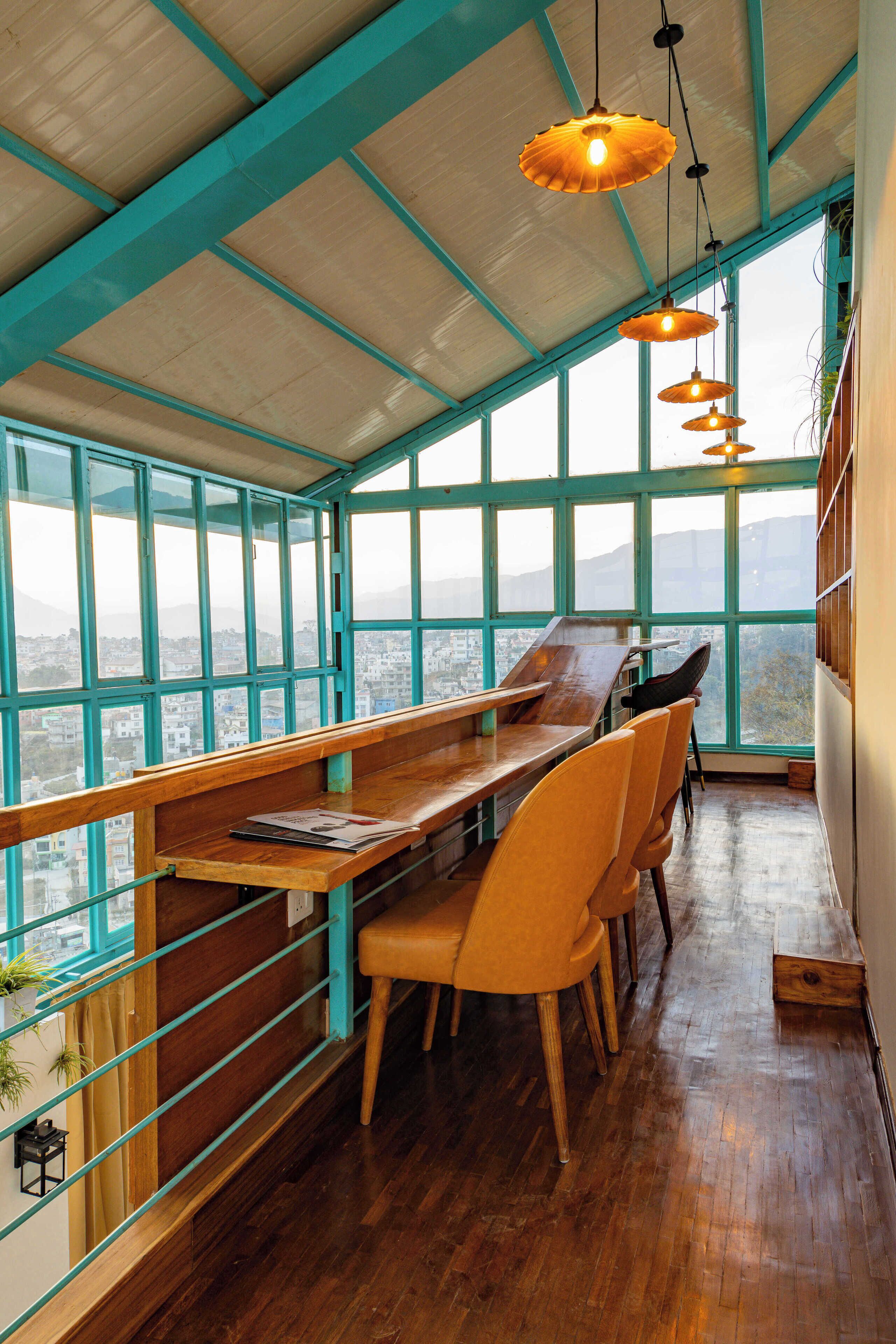 A high-ceilinged room with wooden benches and chairs, large windows offering a city view, and pendant lighting.