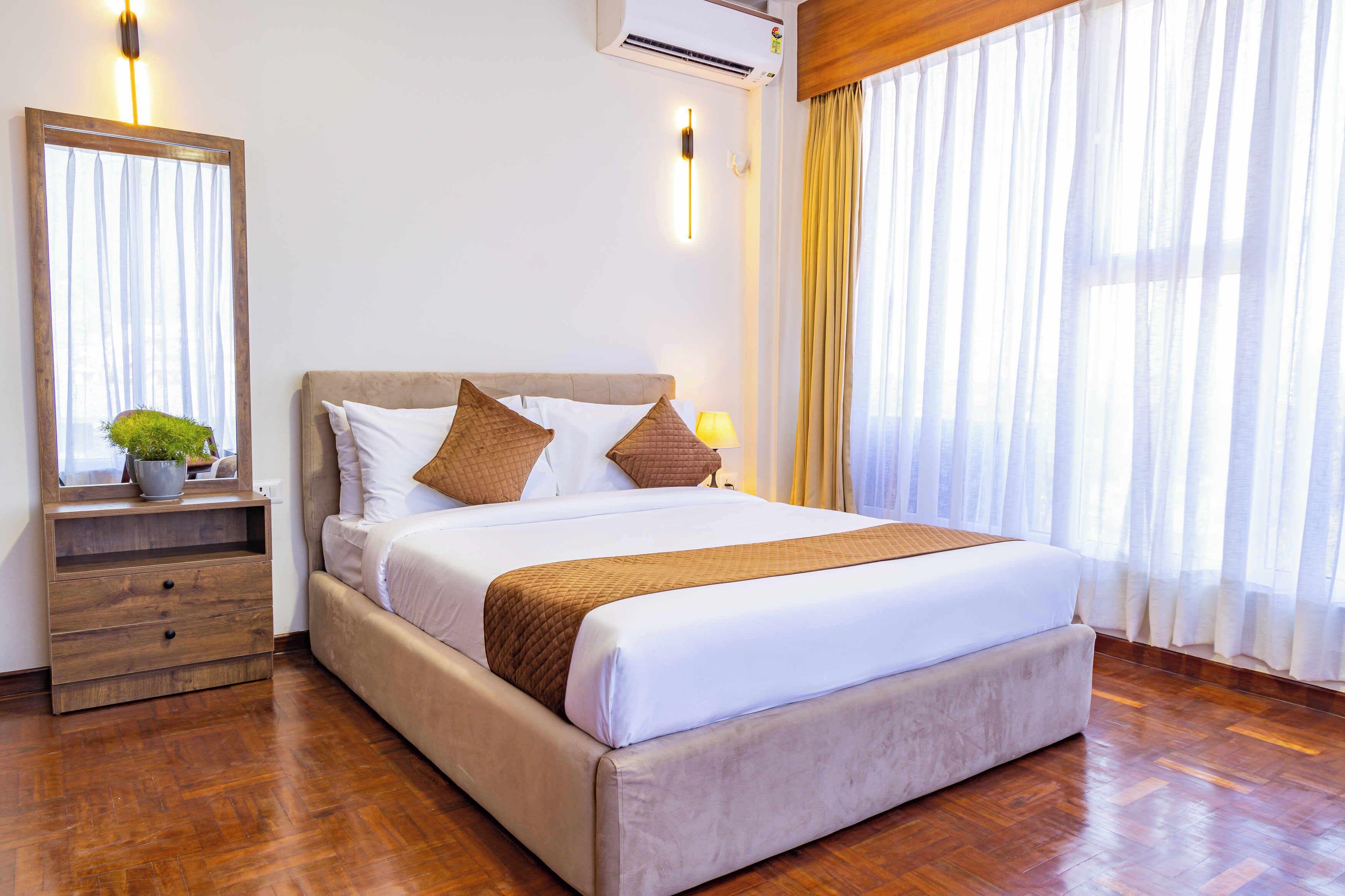 Una habitación de hotel ordenada con una cama grande, una mesita de noche con lámpara, un espejo y una planta en maceta.