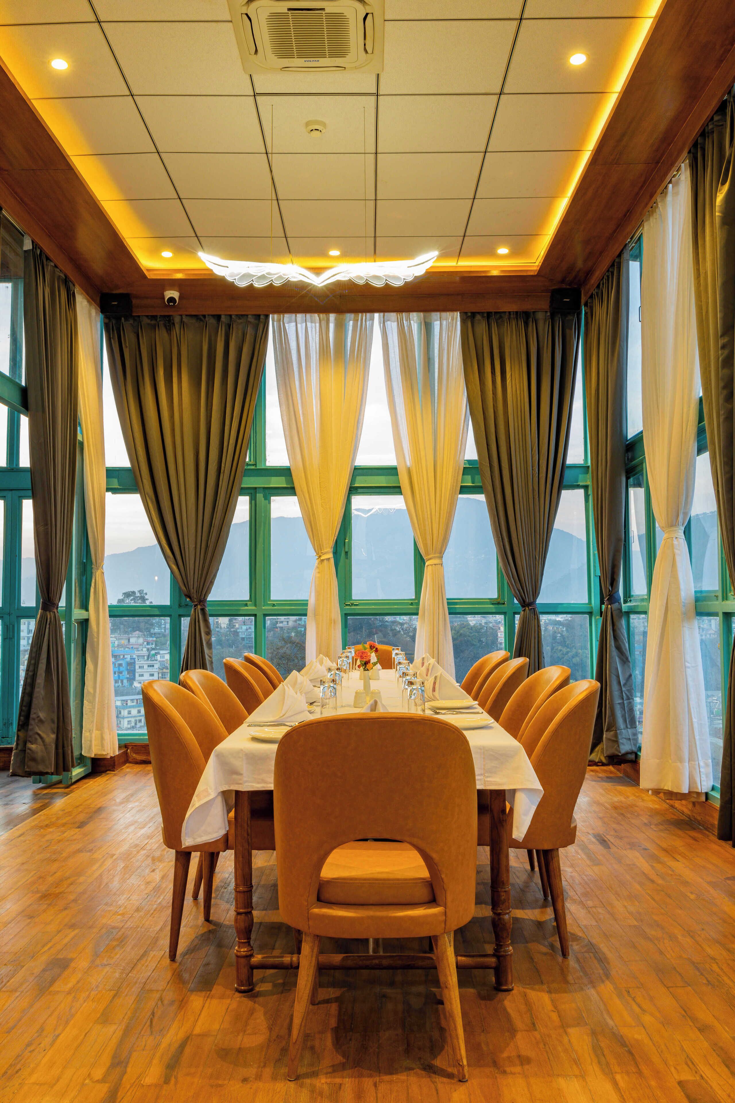 A dining area with a long table set for six, surrounded by chairs, large windows with curtains, and a view of a cityscape.