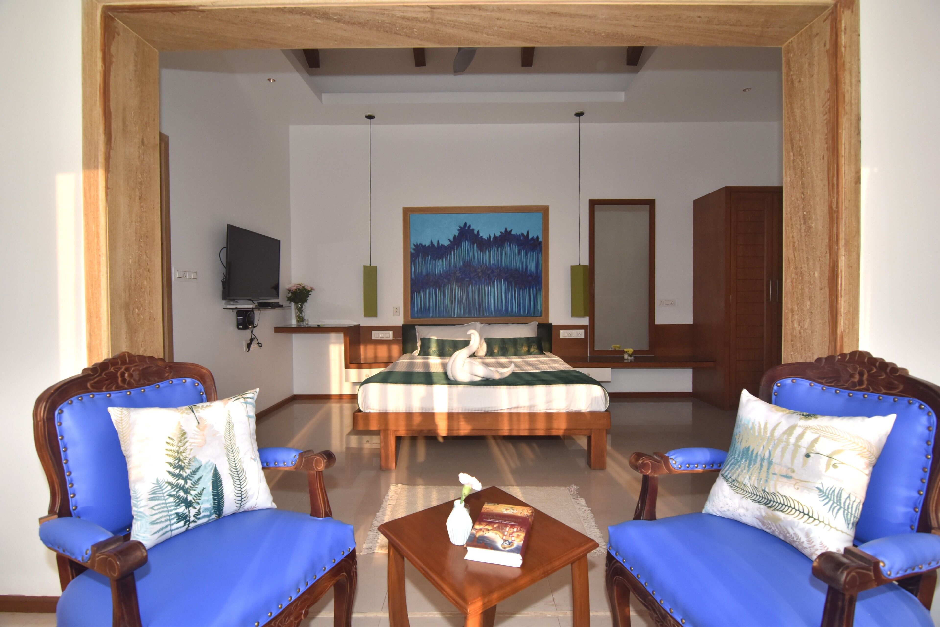 Habitación de hotel con una cama grande, dos sillones tapizados en azul, una mesita con un libro y un jarrón, un televisor y un cuadro en la pared.
