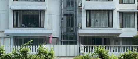 Un edificio residencial de varios pisos con un ascensor central, cerca viva y un seto blanco.