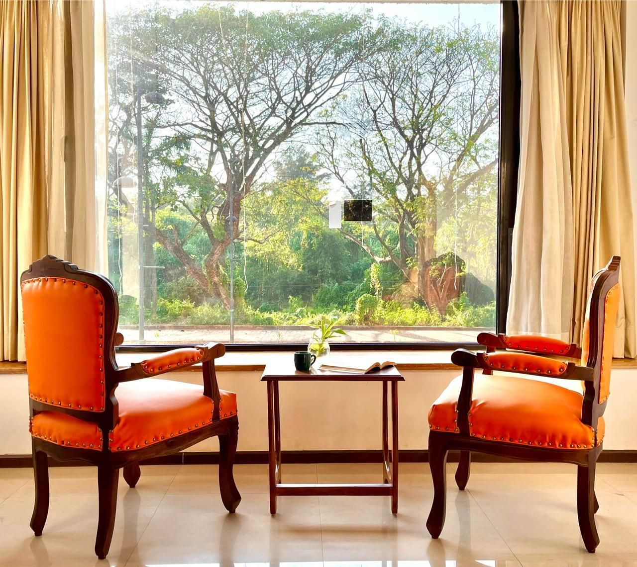 Two orange upholstered chairs with wooden frames and a small table with a plant, positioned in front of a large window with curtains, overlooking a lush green landscape.