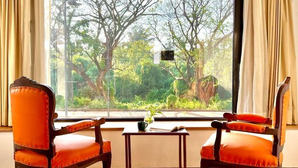 Two orange upholstered chairs with wooden frames and a small table with a plant, positioned in front of a large window with curtains, overlooking a lush green landscape. - Beach Paradise Miramar Goa (Panaji)