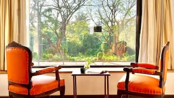 Two orange upholstered chairs with wooden frames and a small table with a plant, positioned in front of a large window with curtains, overlooking a lush green landscape.
