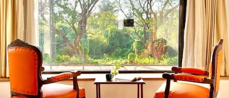 Two orange upholstered chairs with wooden frames and a small table with a plant, positioned in front of a large window with curtains, overlooking a lush green landscape.
