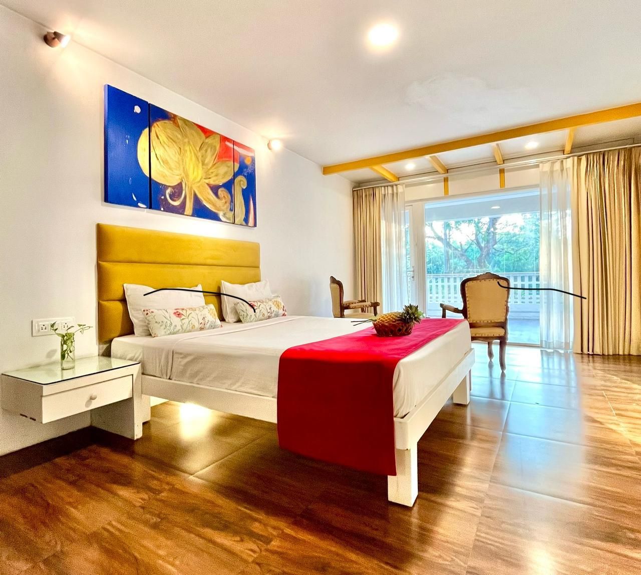 Un dormitorio moderno con suelo de madera, una cama con manta roja, una mesita de noche, una silla y un ventanal con cortinas.