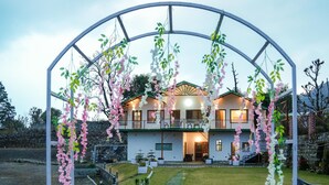 A two-story house with a balcony, a stone wall, and a gravel pathway leading to a white archway adorned with hanging flowers. - Monal meadows (Vijraula, Saikhola)