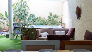 Ein Essbereich mit einem Holztisch, gedeckt für vier Personen, umgeben von einer weißen Wand mit einem dekorativen Wappen. Übertöpfe und im Hintergrund ein Blick auf einen Pool.