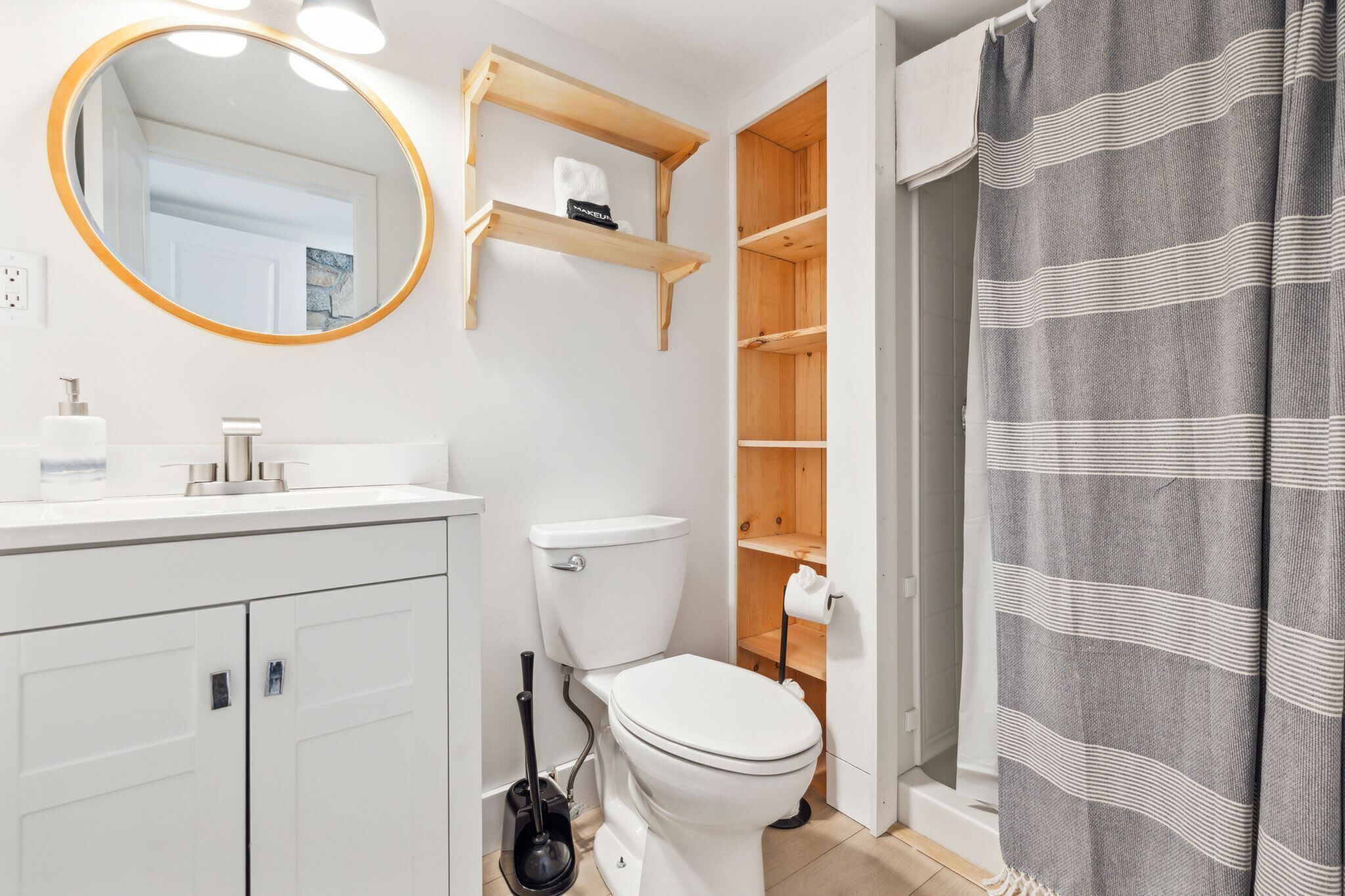 Un baño con un inodoro blanco, un lavabo con espejo, estantes de madera sobre el inodoro y una cortina de ducha.