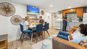 A modern living room with a dining area, a kitchen, and a TV displaying a scenic view. - Across From Beach | Renovated | Parking (Hampton)