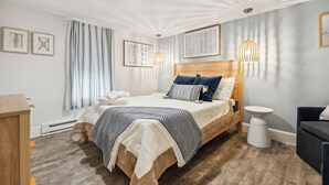 A modern bedroom with a wooden headboard, a large bed with a patterned bedspread, a bedside table, a small stool, and a dark-colored armchair. - Across From Beach | Renovated | Parking (Hampton)
