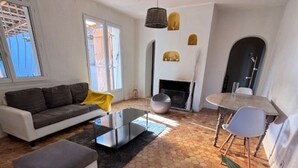 Un salon moderne avec un canapé, une table basse et un coin repas comprenant une table et des chaises. On aperçoit une cheminée et une grande plante au premier plan.