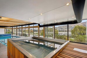Un área de piscina interior moderna con cerramiento de vidrio, terraza de madera y grandes ventanales que ofrecen una vista panorámica.