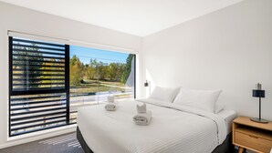 Ett modernt hotellrum med en stor säng, ett träbord som sängbord och utsikt över en väg och träd genom stora fönster som sträcker sig från golv till tak.
