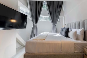 Une chambre moderne avec un grand lit, des rideaux gris, un téléviseur fixé au mur et une vue sur la verdure à l’extérieur.