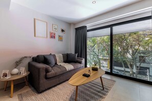Una sala de estar moderna con un sofá, una mesa de centro, una mesita auxiliar y un ventanal con vistas a árboles y edificios.