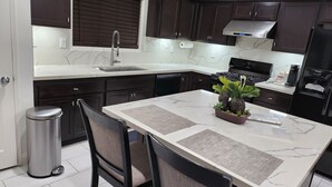 Una cocina moderna con armarios oscuros, encimera de mármol, pileta de acero inoxidable, estufa y refrigerador. Hay un bote de basura y dos sillas en la mesa de comedor.