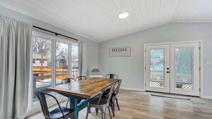 Un comedor con una mesa de madera y sillas de metal, ventanas amplias con cortinas, un letrero que dice 'HOME' en la pared y una puerta con paneles de vidrio.