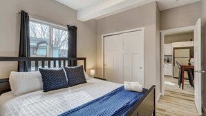 Ein Schlafzimmer mit einem großen Bett, einem Nachttisch, einem Fenster mit Vorhängen, einer Tür und Blick auf einen Küchenbereich.