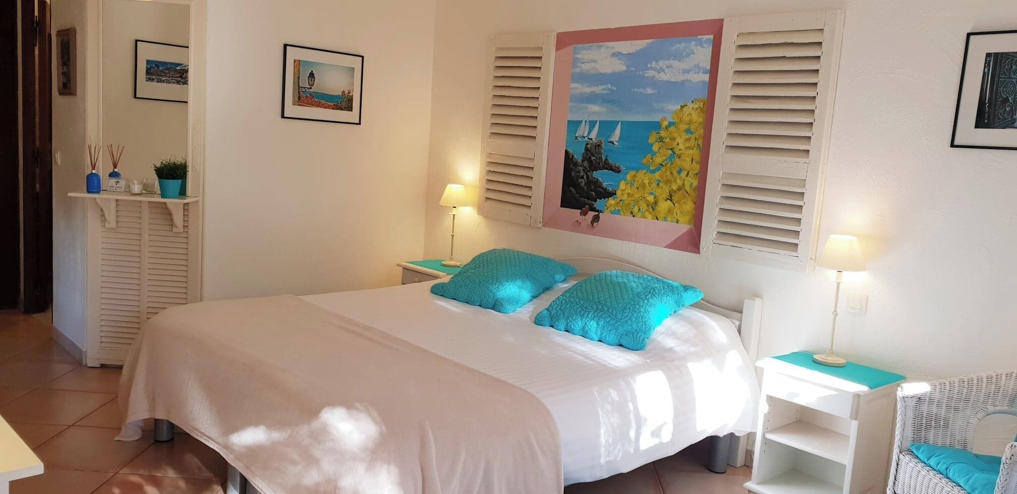 Une chambre à coucher bien rangée, avec un lit, deux tables de chevet et une petite commode. On y trouve deux tableaux encadrés accrochés au mur, une fenêtre avec des volets et une petite étagère ornée d’objets décoratifs.