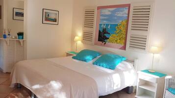 Une chambre à coucher bien rangée, avec un lit, deux tables de chevet et une petite commode. On y trouve deux tableaux encadrés accrochés au mur, une fenêtre avec des volets et une petite étagère ornée d’objets décoratifs.