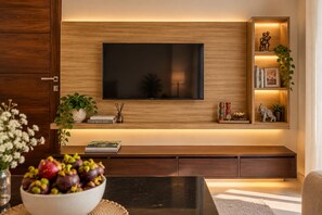 A modern living room with a flat-screen TV mounted on a wooden wall, a bookshelf with decorative items, and a bowl of fruit on a table. - Lake Villas Premium Kandy (Kandy)