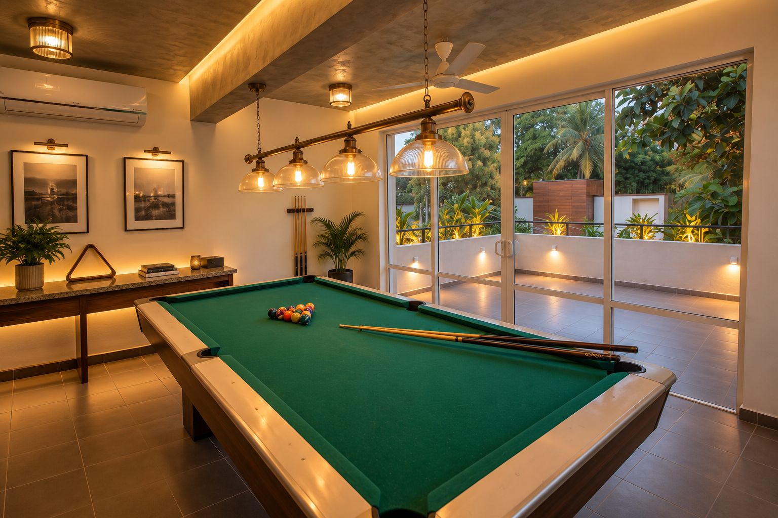 A pool table with a green felt surface, surrounded by a modern interior with a ceiling fan, pendant lighting, and a view of a lush outdoor balcony.