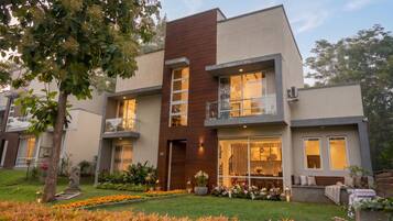 A modern two-story residential building with a balcony, large windows, and a well-maintained garden.