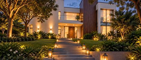 A modern house with a staircase leading to the entrance, surrounded by lush greenery and illuminated by outdoor lights.