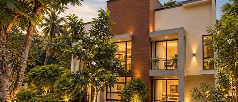 A modern two-story building with large windows and balconies, surrounded by lush greenery and palm trees.