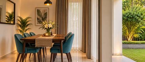 A dining area with a wooden table, blue chairs, a hanging light fixture, and a view of a garden through large windows.