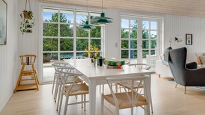 Une pièce spacieuse avec une table à manger blanche, des chaises blanches, un canapé gris et de grandes fenêtres donnant sur un paysage verdoyant.