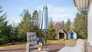 Une terrasse en bois avec des meubles en osier, un parasol bleu et deux petites cabanes en arrière-plan.