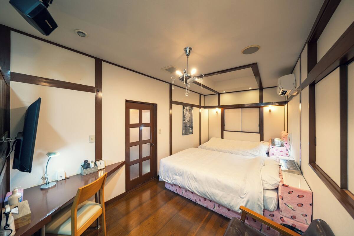 A hotel room with a wooden desk, chair, two beds, a desk lamp, a wall-mounted TV, and a ceiling light fixture.