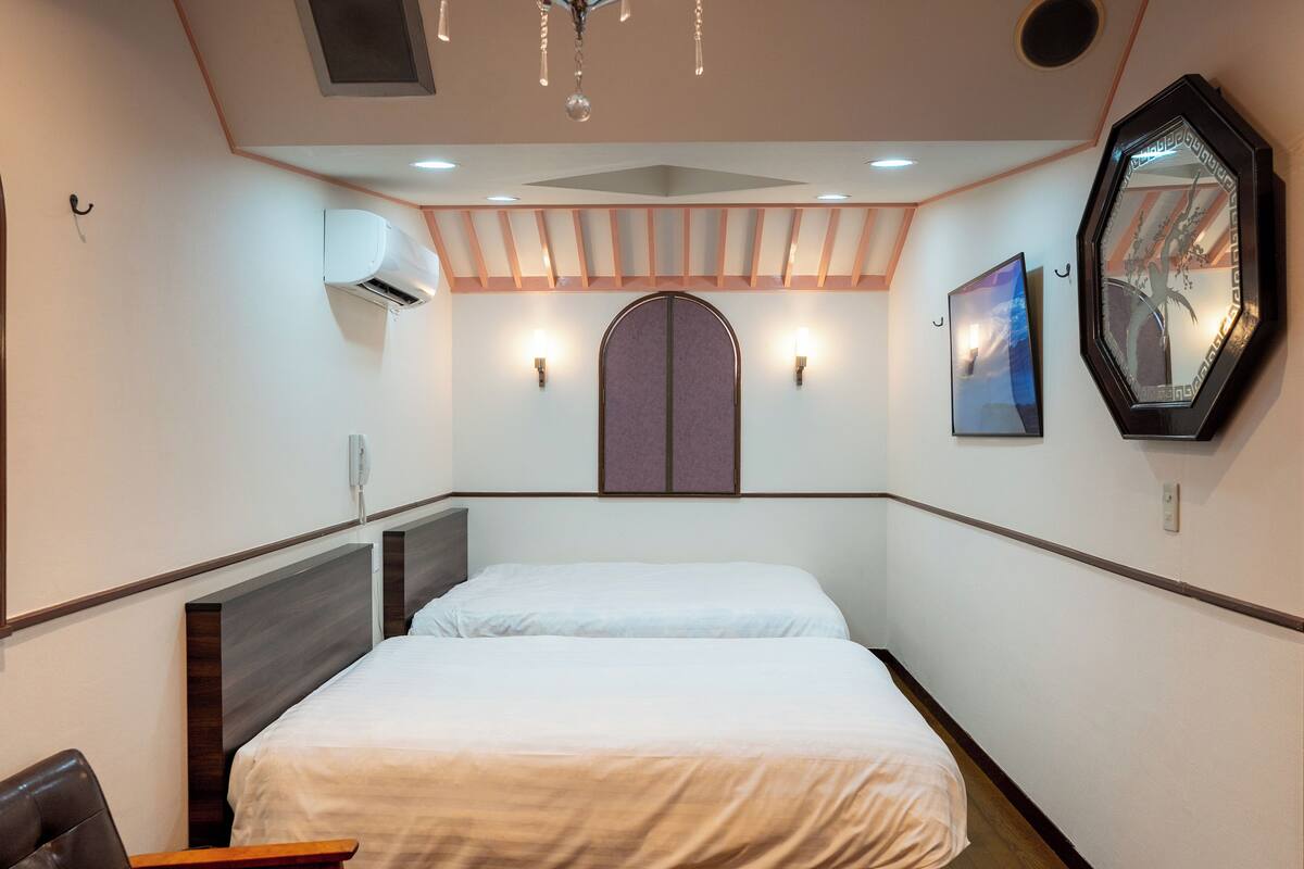 A hotel room with a bed, a chair, an air conditioning unit, a wall-mounted mirror, and two wall sconces.