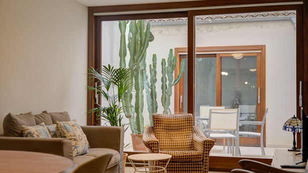 A living room with a sofa, armchairs, a small table, and a large window with a view of cacti and a door. - Comtat Muro 1 by Renallorca (Muro)