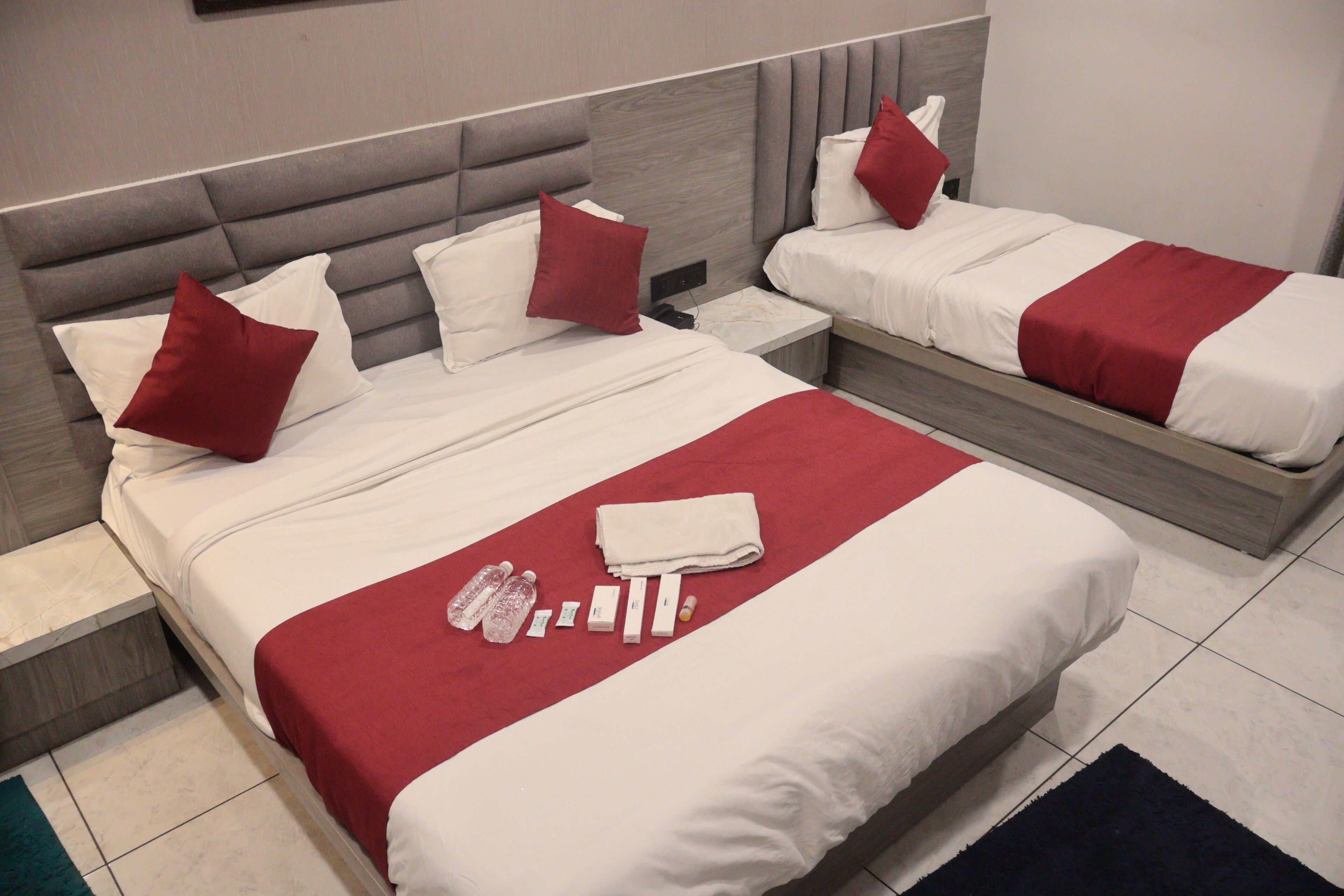 Ein Hotelzimmer mit zwei Betten, jeweils mit weißer Bettwäsche und roten Akzenten. Auf den Betten liegen Pflegeprodukte, ein Nachttisch und ein dunkler Teppich liegt auf dem Boden.