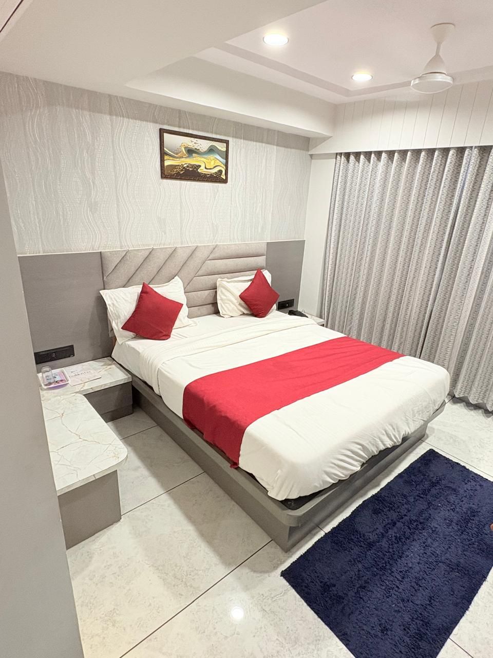 A modern hotel room with a bed, nightstands, a marble-tiled floor, a ceiling fan, and a large window with curtains.