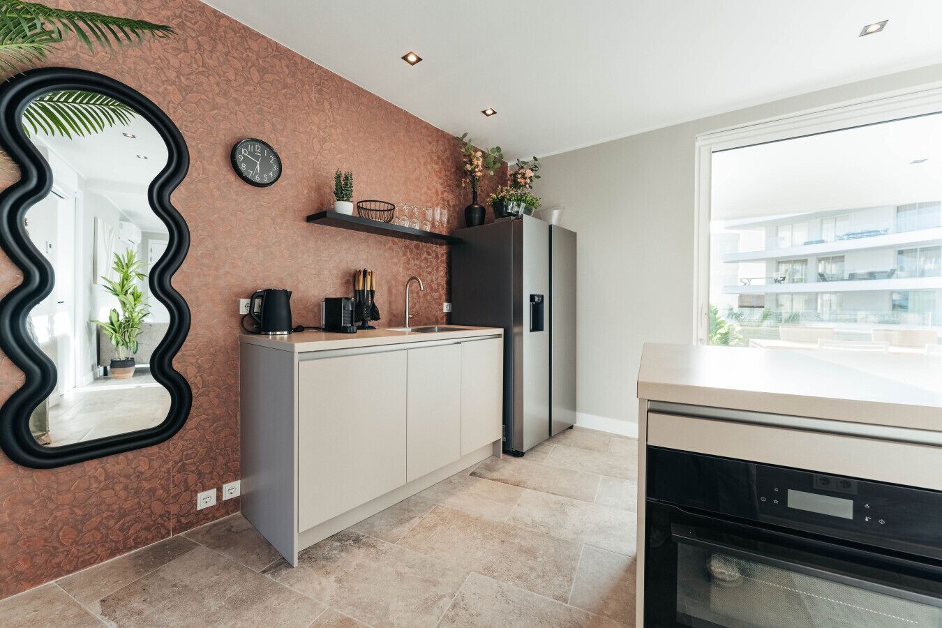 A modern kitchen with a stone backsplash, a black wavy mirror, a clock, and a refrigerator.