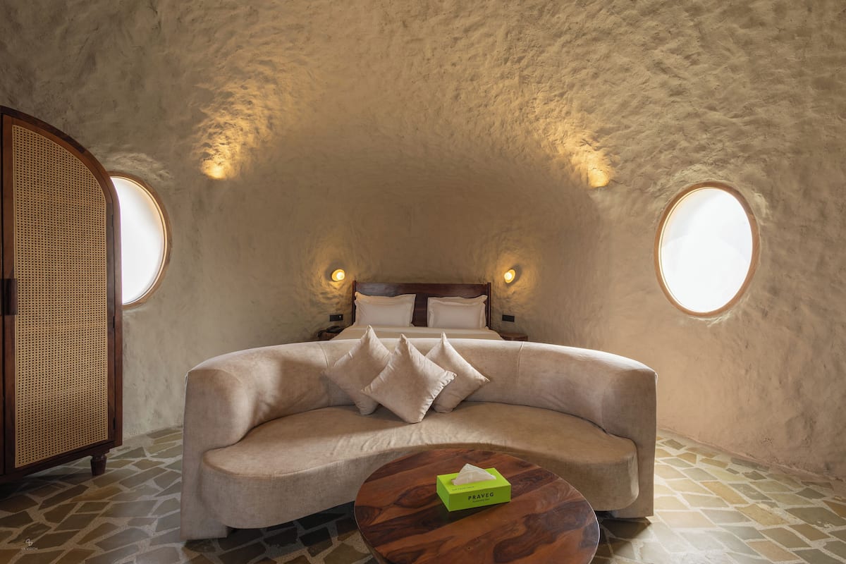 A cozy, circular room with a bed, two windows, a wooden table with a tissue box, and a rattan chair.